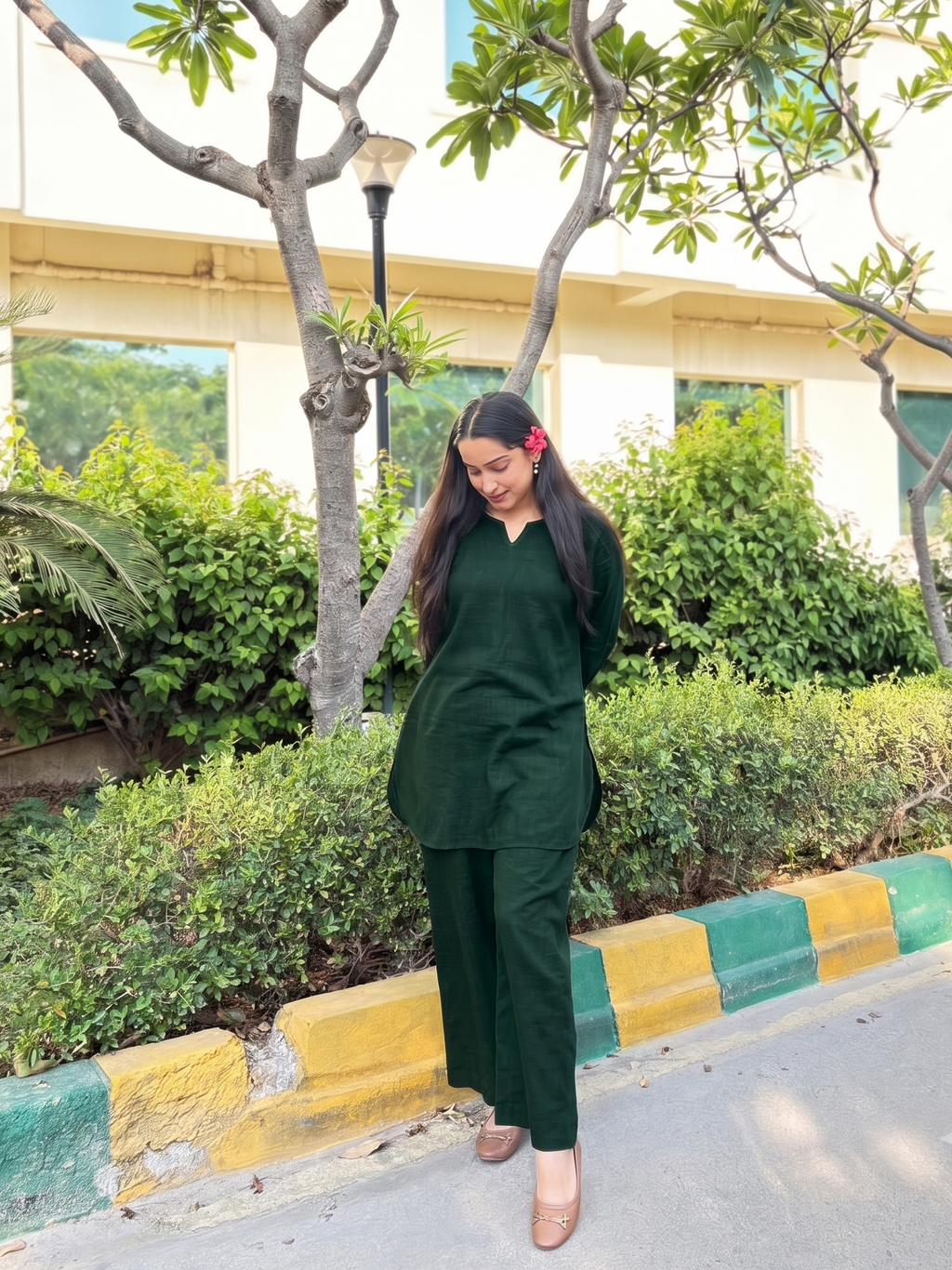 A woman wearing a dark green two-piece kurta set stands outdoors on a paved walkway next to a curb with green and yellow stripes, with trees and a building in the background.