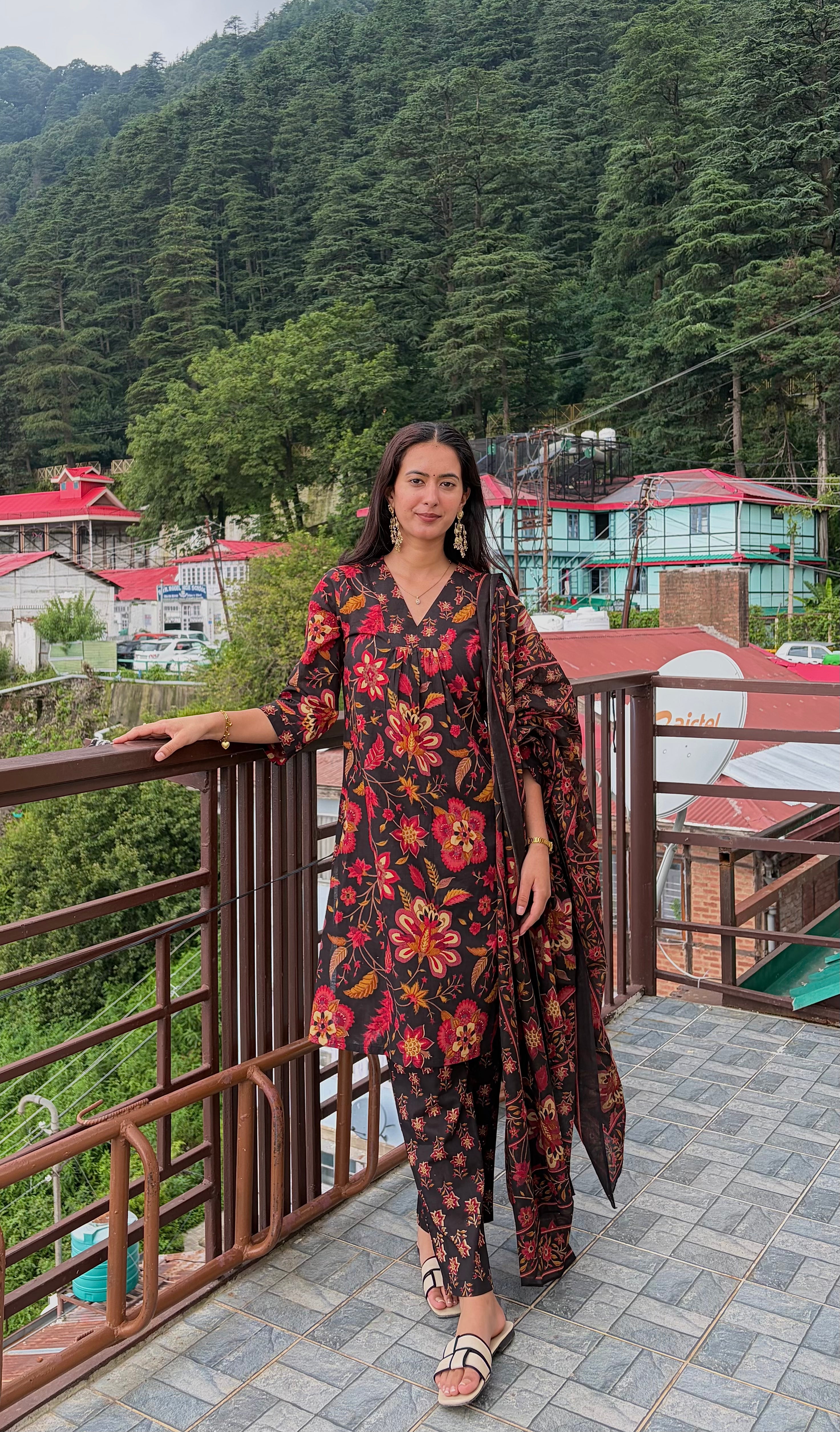 A woman models a dark floral printed kurta set with matching pants and a dupatta on a balcony overlooking a lush green mountain village.