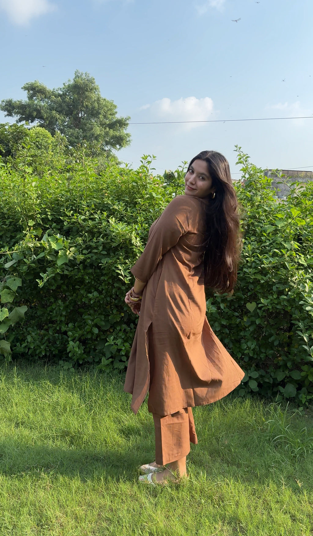 A woman wearing a rust-brown long kurta and matching pants stands outdoors on a grassy lawn in front of a green hedge under a blue sky.