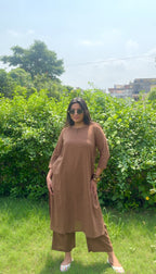 A woman wearing a brown long-sleeve kurta and matching cropped pants stands outdoors in a grassy area with green bushes and trees in the background.