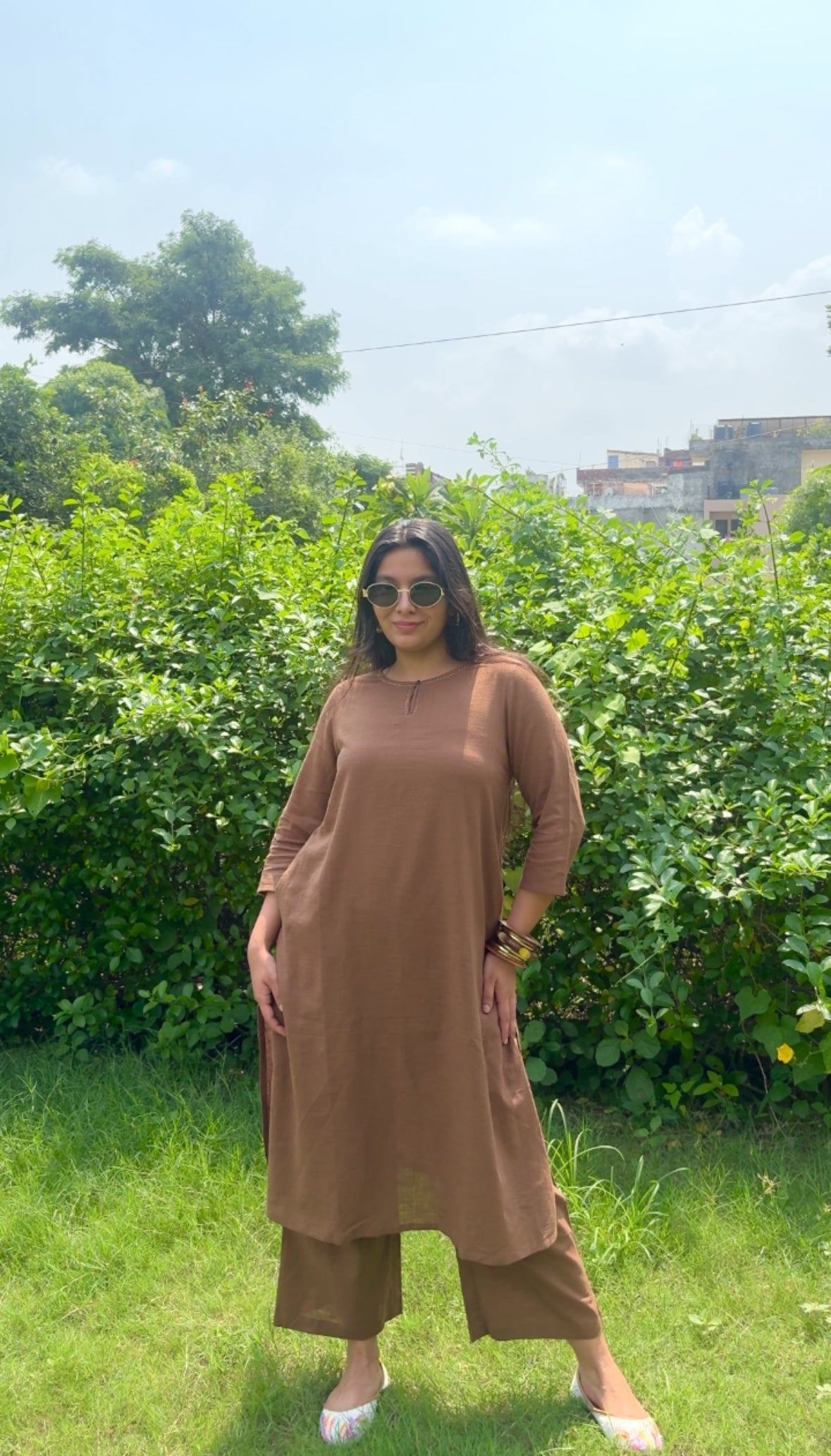 A woman wearing a brown long-sleeve kurta and matching cropped pants stands outdoors in a grassy area with green bushes and trees in the background.