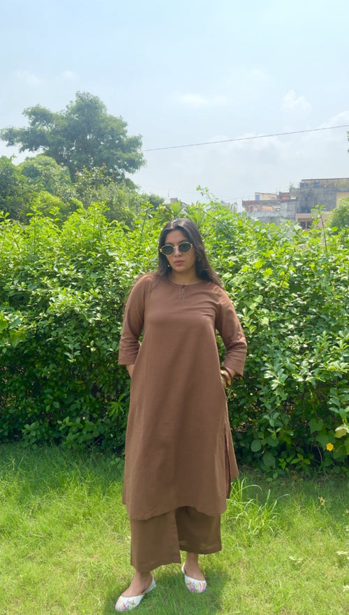 A woman wearing a brown long-sleeve kurta and matching wide-leg pants stands outdoors on a grassy lawn with a green hedge and trees in the background.