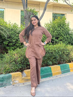 Woman in a brown outfit standing outdoors with greenery and a building in the background